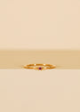 The Pinky Ring - Pink, featuring a small pink cubic zirconia on a delicate gold band, is displayed upright against a minimalist beige and cream backdrop.