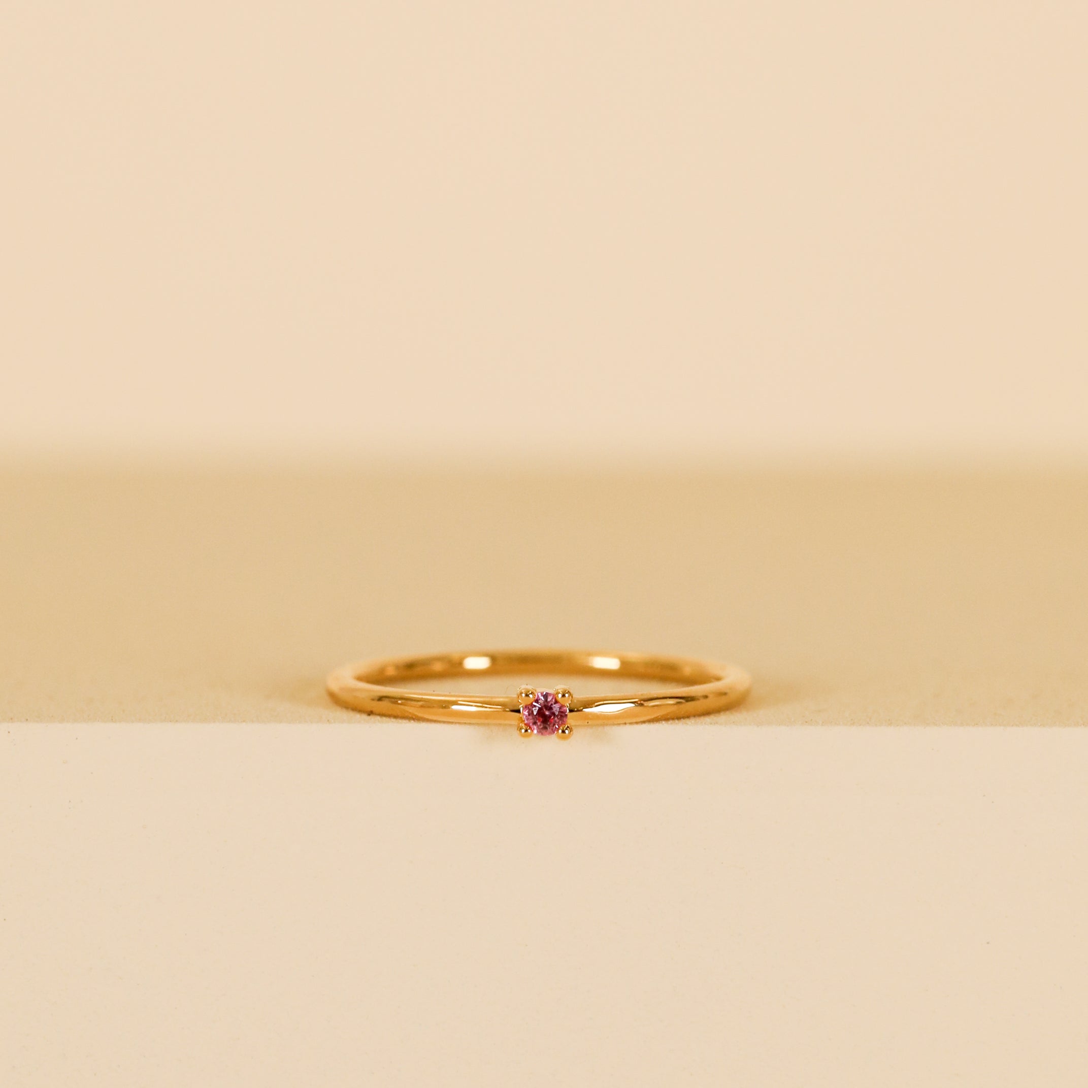 The Pinky Ring - Pink, featuring a small pink cubic zirconia on a delicate gold band, is displayed upright against a minimalist beige and cream backdrop.