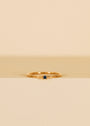 The Pinky Ring - Blue is a delicate 18kt gold ring with a small dark blue cubic zirconia at the center, shown on a beige and cream background.