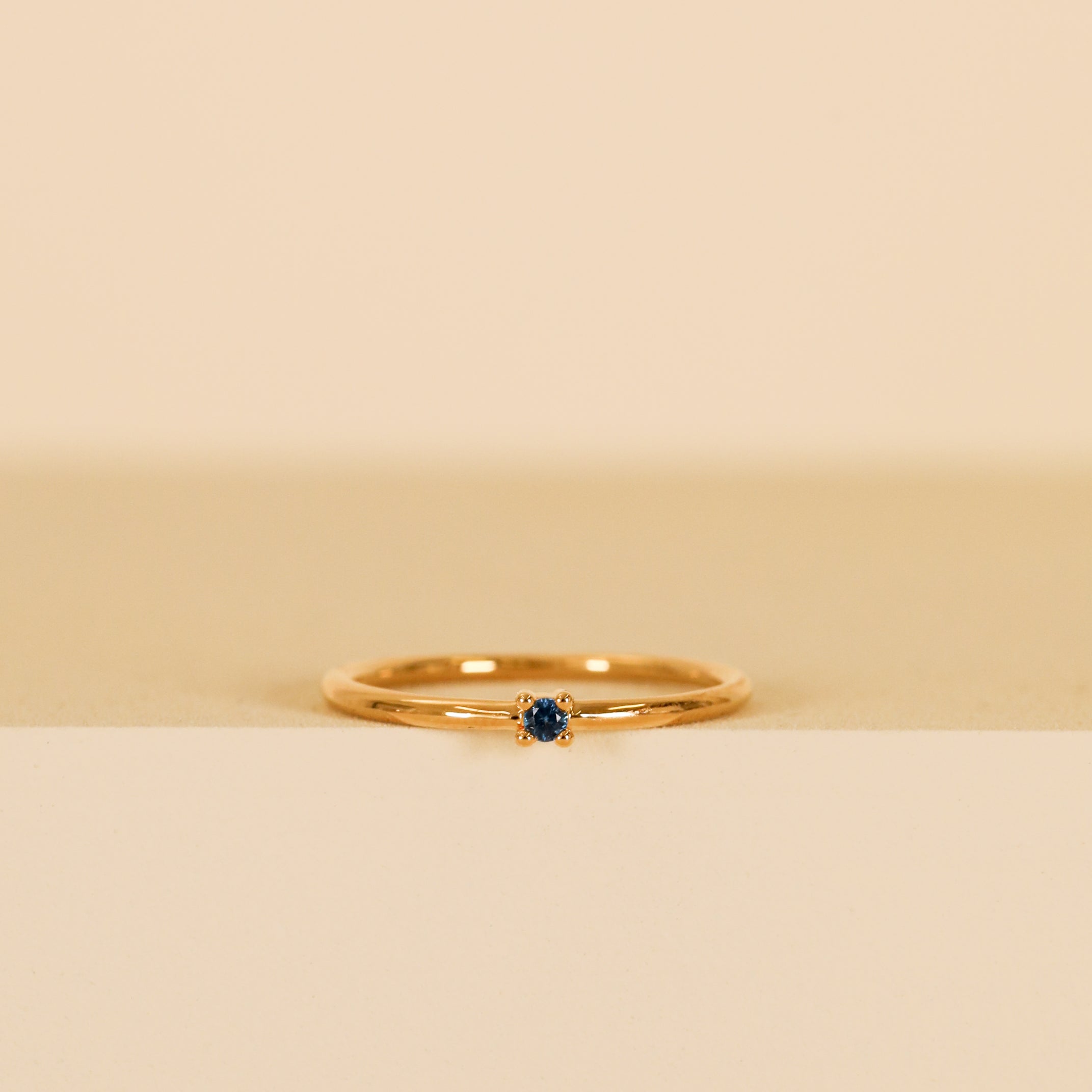 The Pinky Ring - Blue is a delicate 18kt gold ring with a small dark blue cubic zirconia at the center, shown on a beige and cream background.