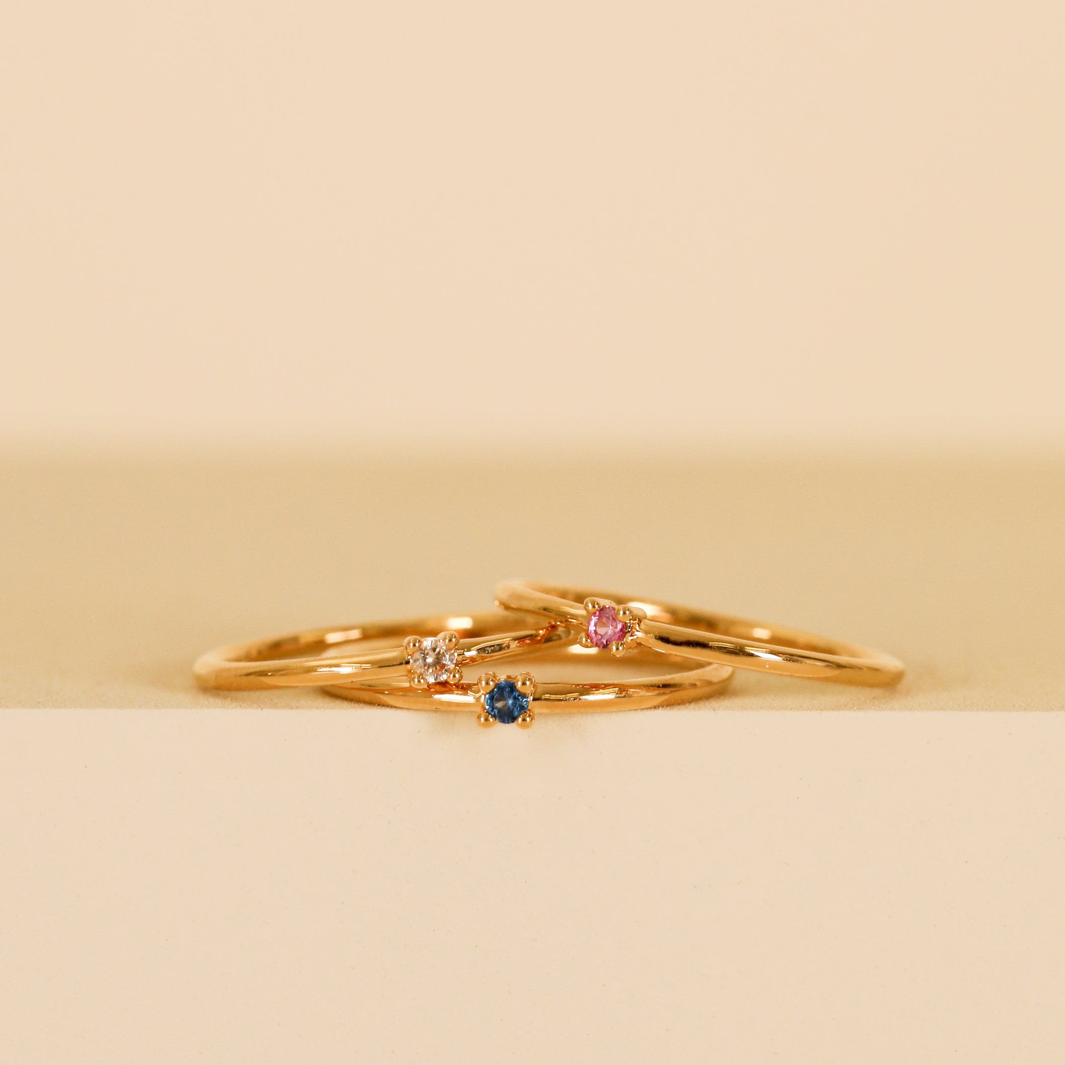 The Pinky Ring - Pink features three dainty gold bands with colorful gemstones—blue, pink cubic zirconia, and clear—stacked against a soft beige background, perfect for wearing solo on your pinky or stacking together.