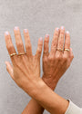 Two hands with light pink manicured nails, wearing several 18kt gold rings including the Pinky Ring - Pink, are crossed at the wrists against a light gray textured background.
