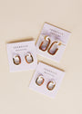 Three pairs of Color Dipped Rectangle Hoop Earrings in brown, featuring gold-tone and enamel geometric hoops on white JaxKelly cards with sterling silver posts and "Wear Good Vibes" slogan, are displayed on a light beige surface.
