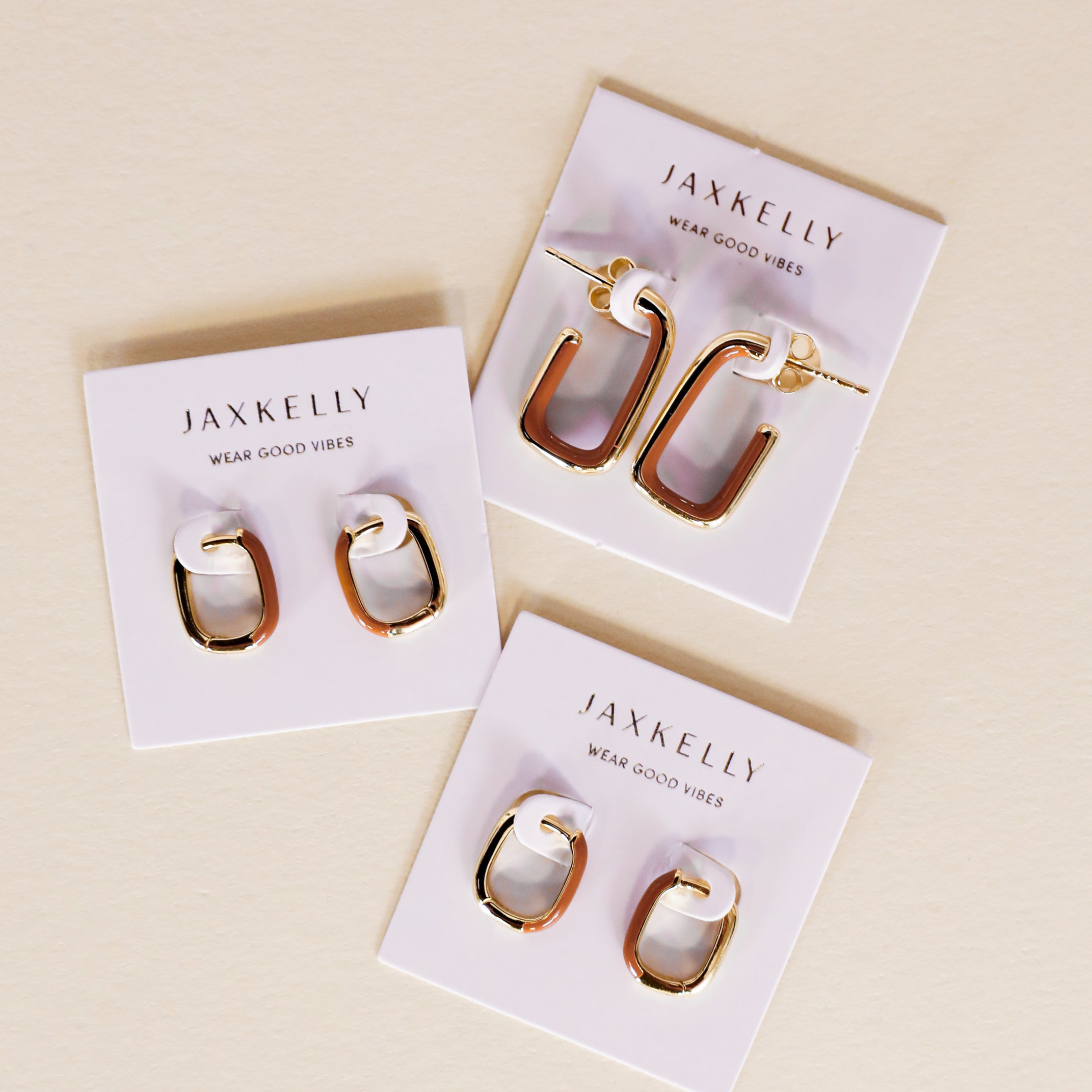 Three pairs of Color Dipped Rectangle Hoop Earrings in brown, featuring gold-tone and enamel geometric hoops on white JaxKelly cards with sterling silver posts and "Wear Good Vibes" slogan, are displayed on a light beige surface.