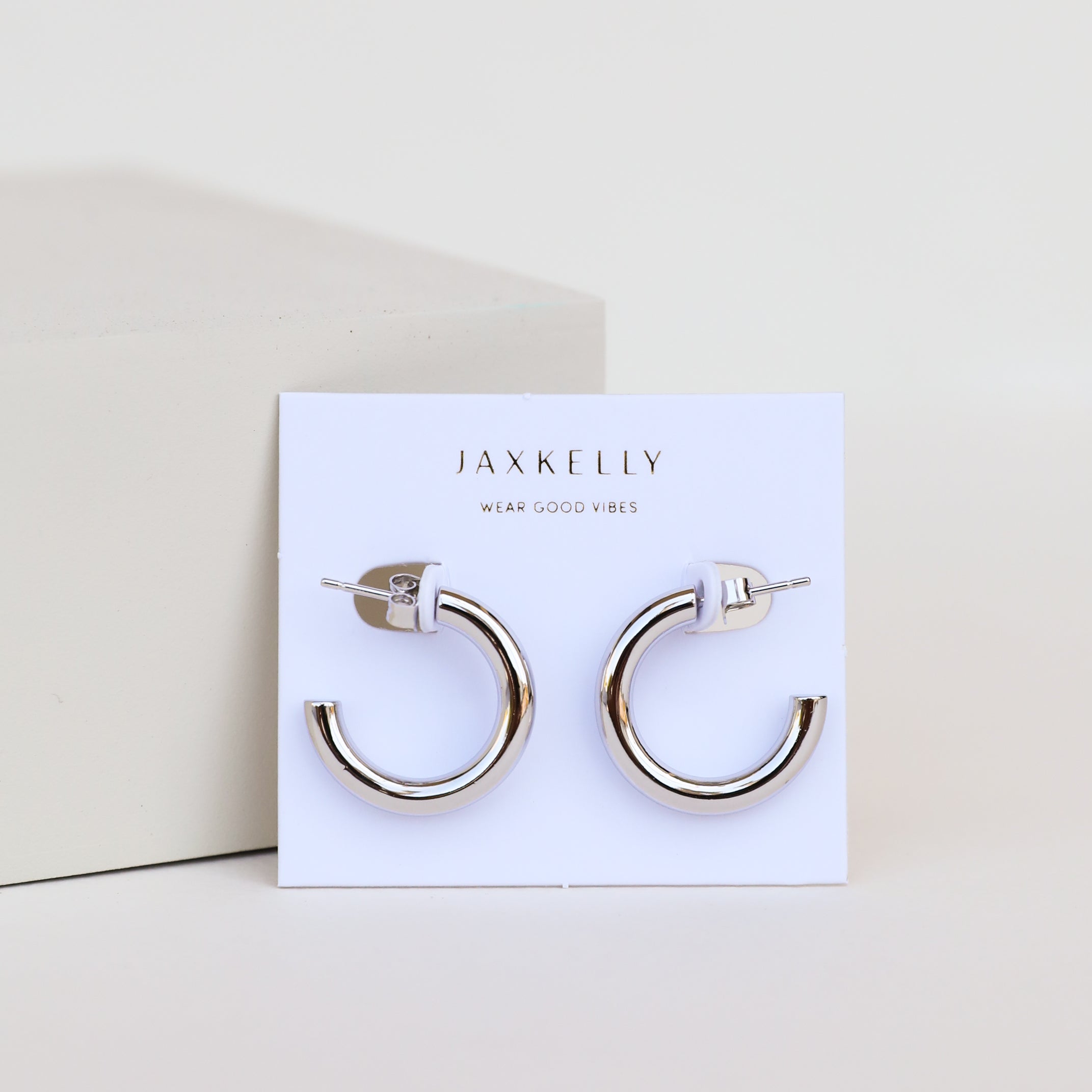 A pair of Silver Hoop - Smooth sterling silver earrings with rhodium plating is displayed on a white JaxKelly card that reads "Wear Good Vibes," set against a minimal pale background.