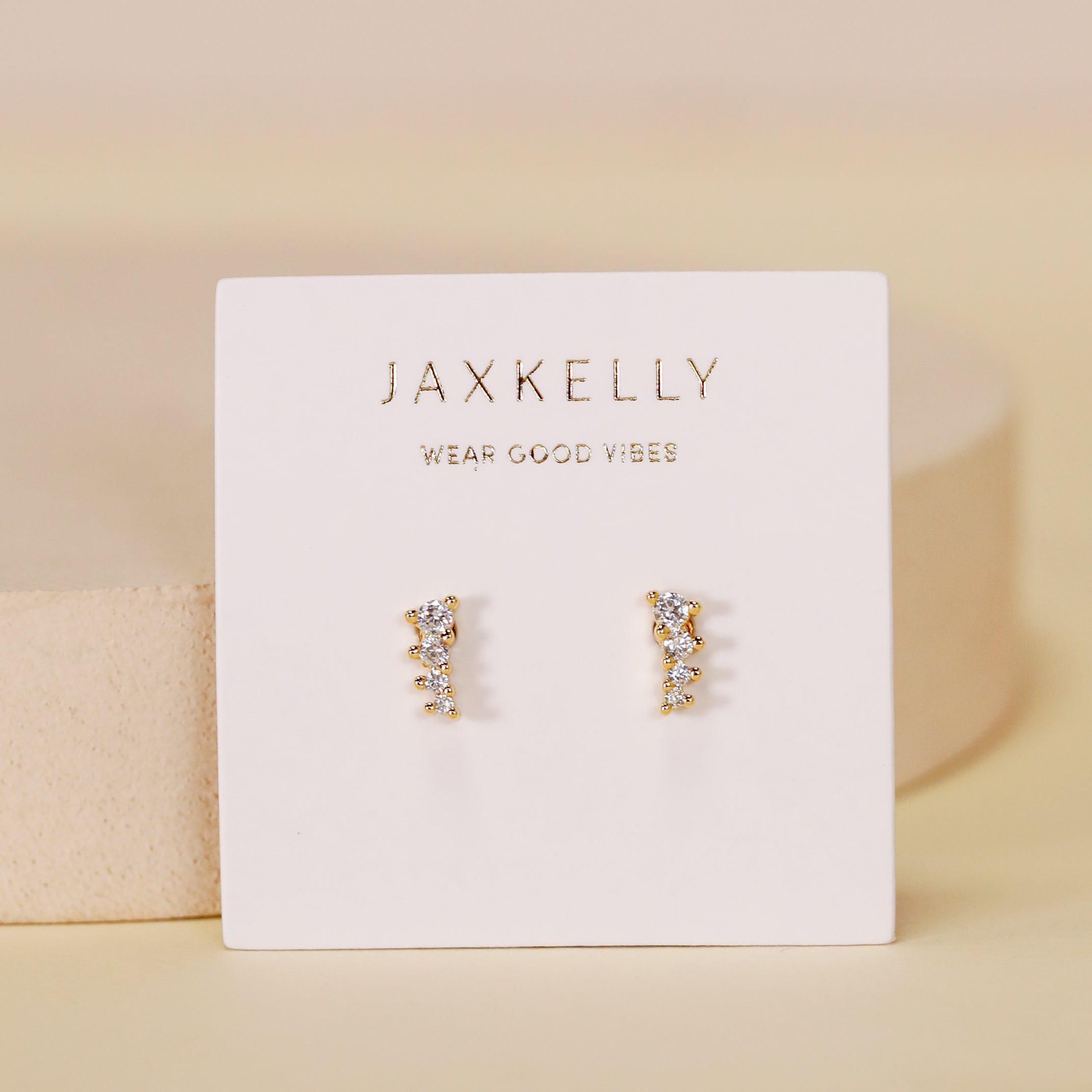 The Mini Crawler - White CZ - Gold Earrings are shown on a white card with "JaxKelly" and "Wear Good Vibes" in gold lettering, set against a soft beige background.