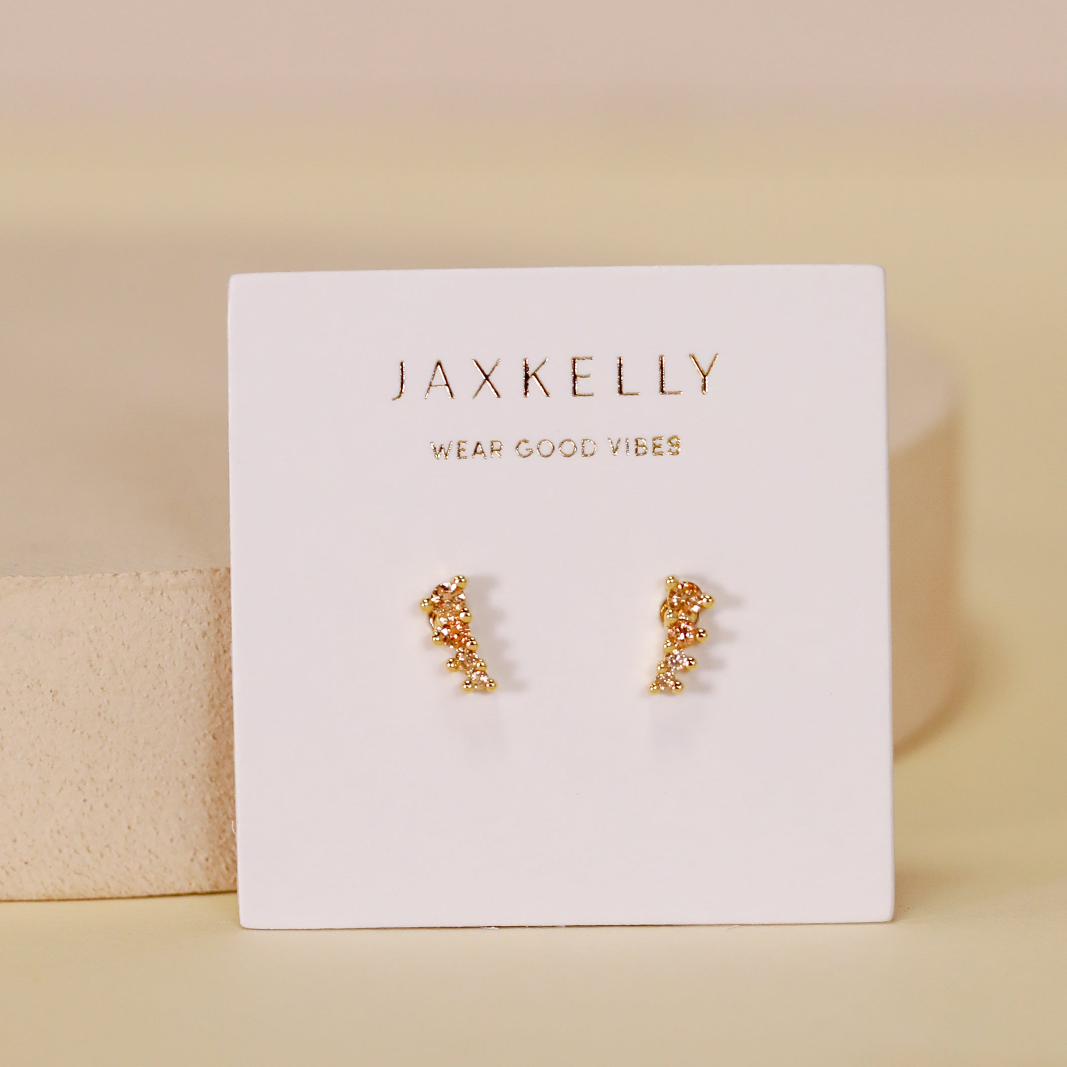 The Mini Crawler - Champagne CZ - Gold Earrings, featuring small gold plated studs with sparkling champagne CZ stones, are displayed on a JaxKelly Wear Good Vibes card against a soft beige, minimal background.
