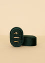 A Small Oval Velvet Jewelry Box in dark green holds three delicate gold rings—one with a row of small stones—elegantly displayed against a plain beige background.
