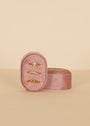 A Small Oval Velvet Jewelry Box in pink velvet holds three delicate gold rings, including one with a small gemstone. The slightly open box is displayed on a neutral beige background.