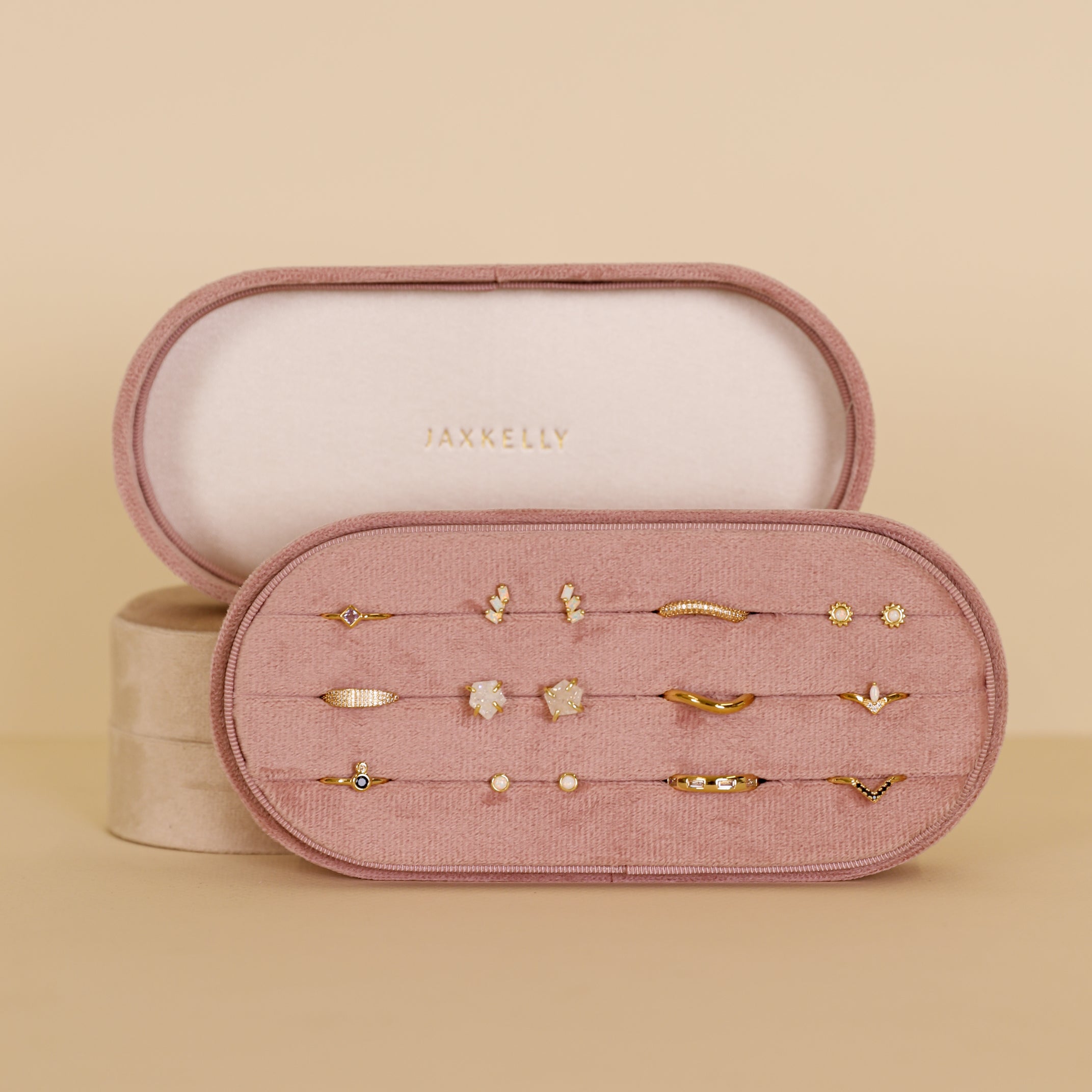 The Large Oval Velvet Jewelry Box in Light Pink by JaxKelly is a travel-friendly organizer that opens to display twelve pairs of small gold earrings—hoops, studs, and more—in neat rows. The inside lid showcases the JaxKelly brand name.