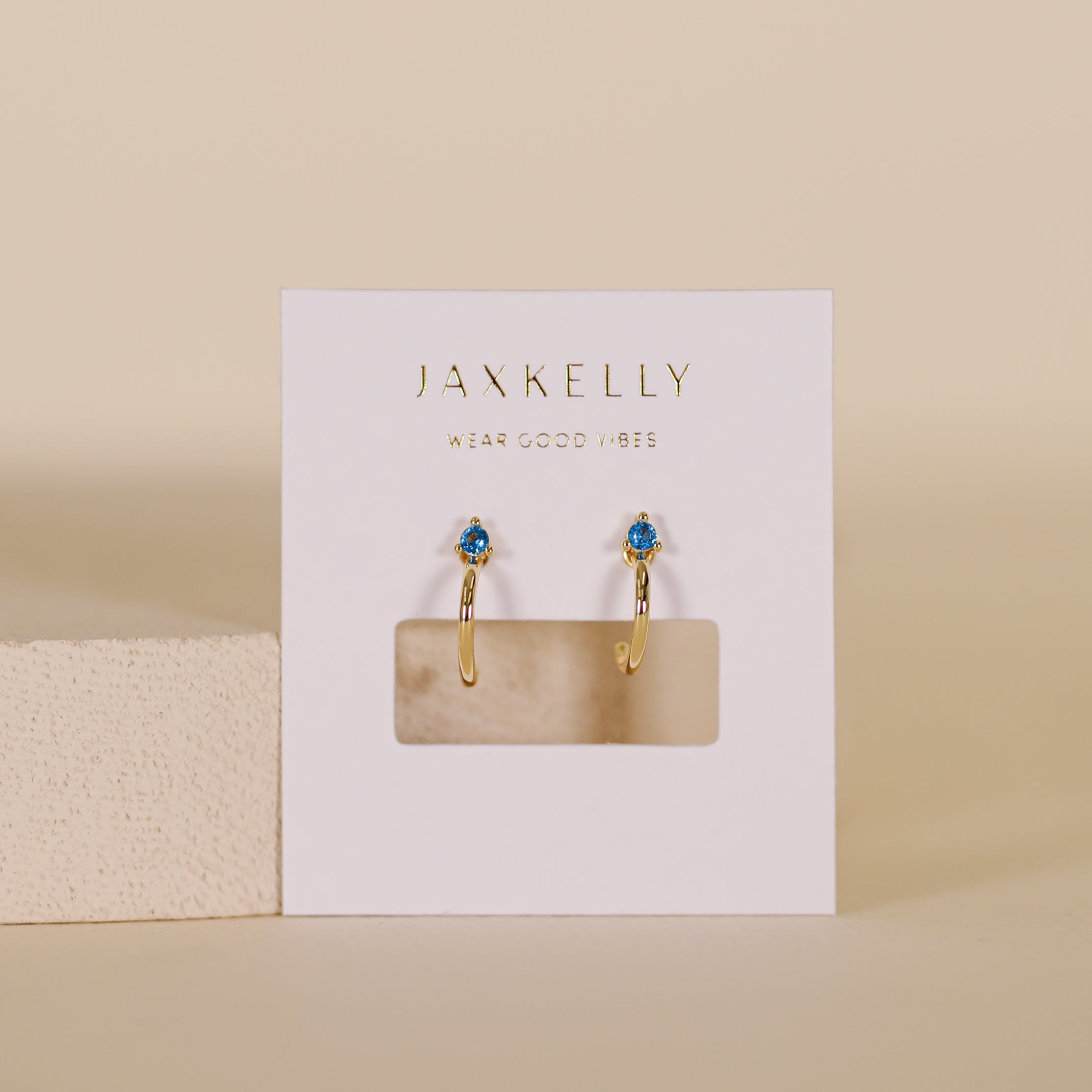 The Open Mini Hoop Earrings - Blue Sapphire feature sapphire CZ stone studs and 18K gold plating, shown on a white JaxKelly card against a neutral beige background.