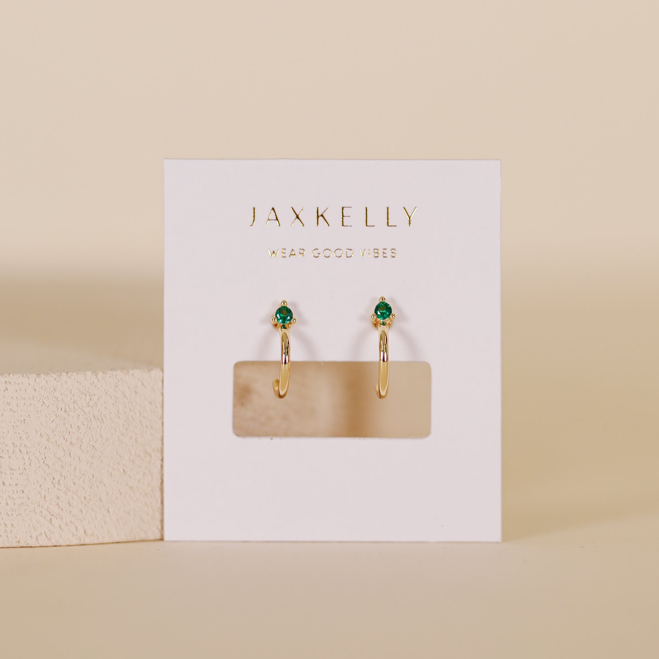 A pair of Open Mini Hoop Earrings - Green Emerald, gold plated with emerald CZ studs, displayed on a white JaxKelly card with gold text against a neutral beige background.