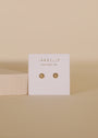 A pair of Petite Daisy Stud gold earrings, displayed on a white JAX KELLY WEAR GOOD VIBES card against a neutral beige background—perfect for adding to your ear stack.