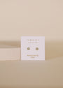 The Simple Stud Earring - Amazonite Gemstone features small round amazonite studs with sterling silver posts on a white JaxKelly card labeled "AMAZONITE hope," set against a beige background with a light textured block to the left.