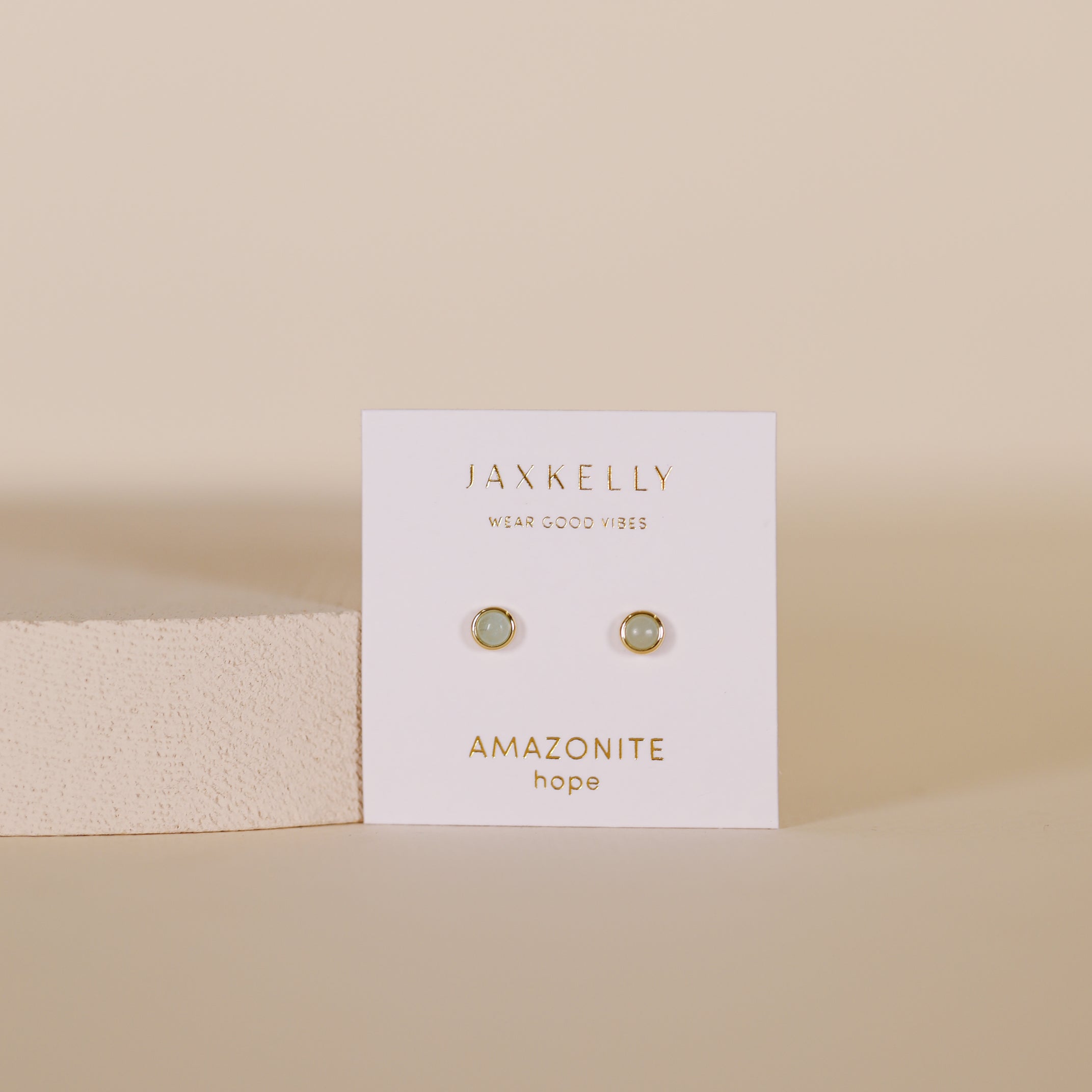The Simple Stud Earring - Amazonite Gemstone features small round amazonite studs with sterling silver posts on a white JaxKelly card labeled "AMAZONITE hope," set against a beige background with a light textured block to the left.