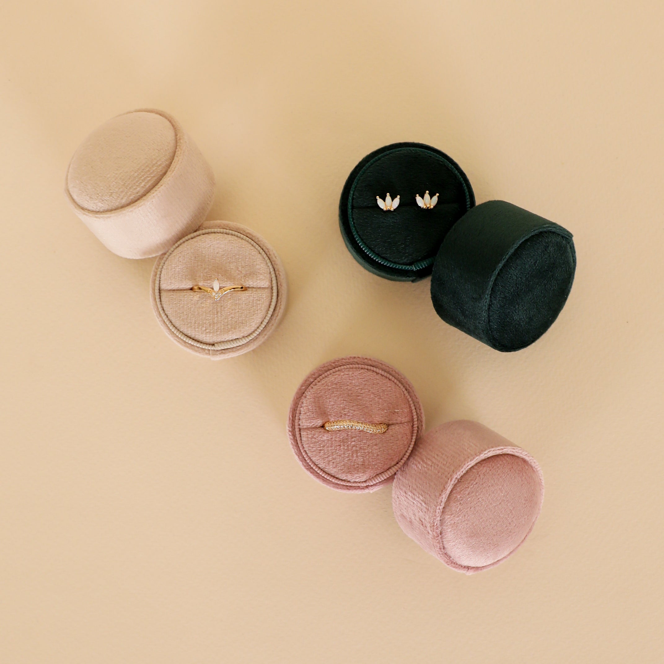 Two JaxKelly Circle Velvet Jewelry Boxes in Light Pink and one in Dark Green hold minimalist earrings and a ring, displayed on a beige surface—perfect for adding to your travel jewelry organizer.