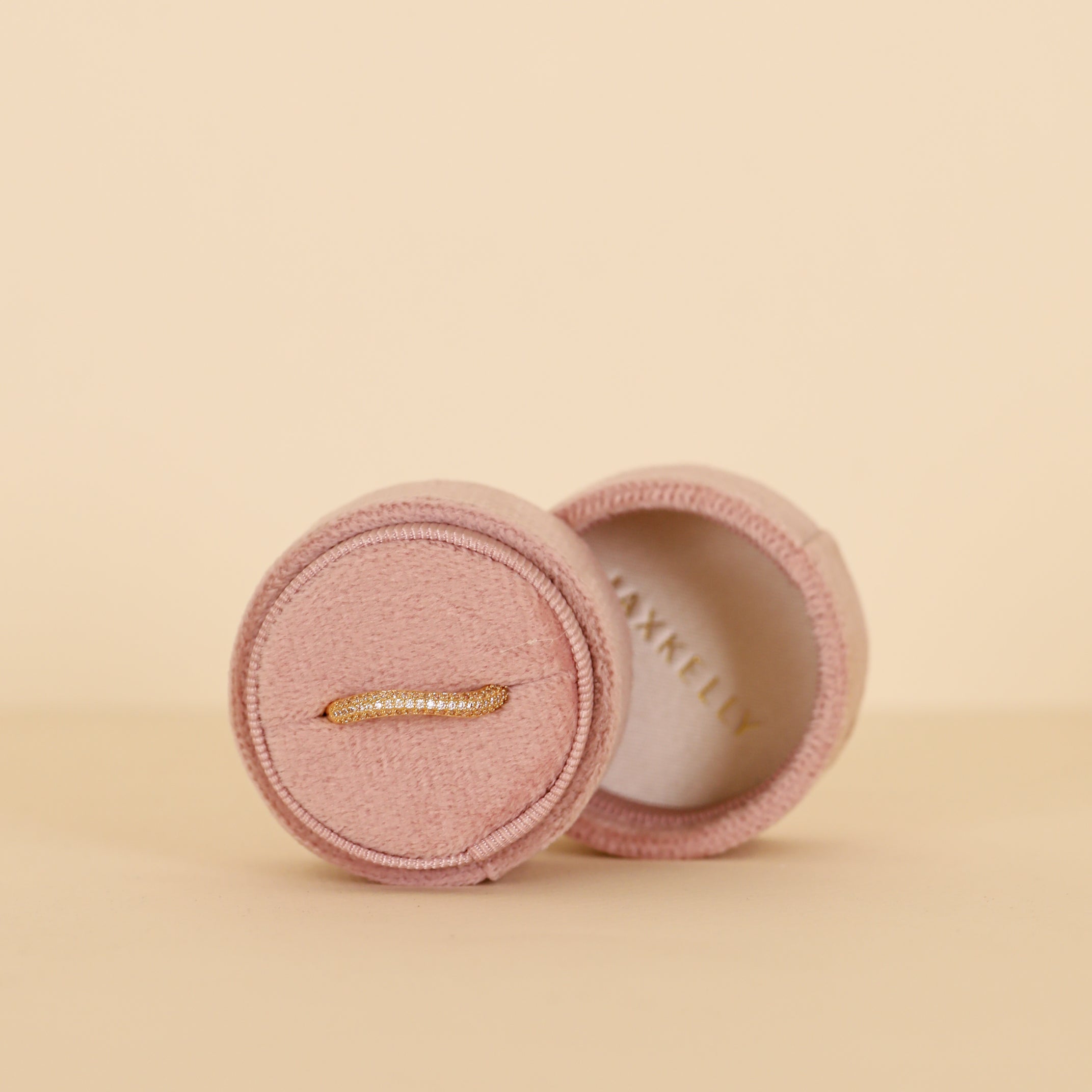 A delicate gold ring with small diamonds is shown in the open Circle Velvet Jewelry Box - Light Pink by JaxKelly, with the lid propped behind it against a soft beige background.