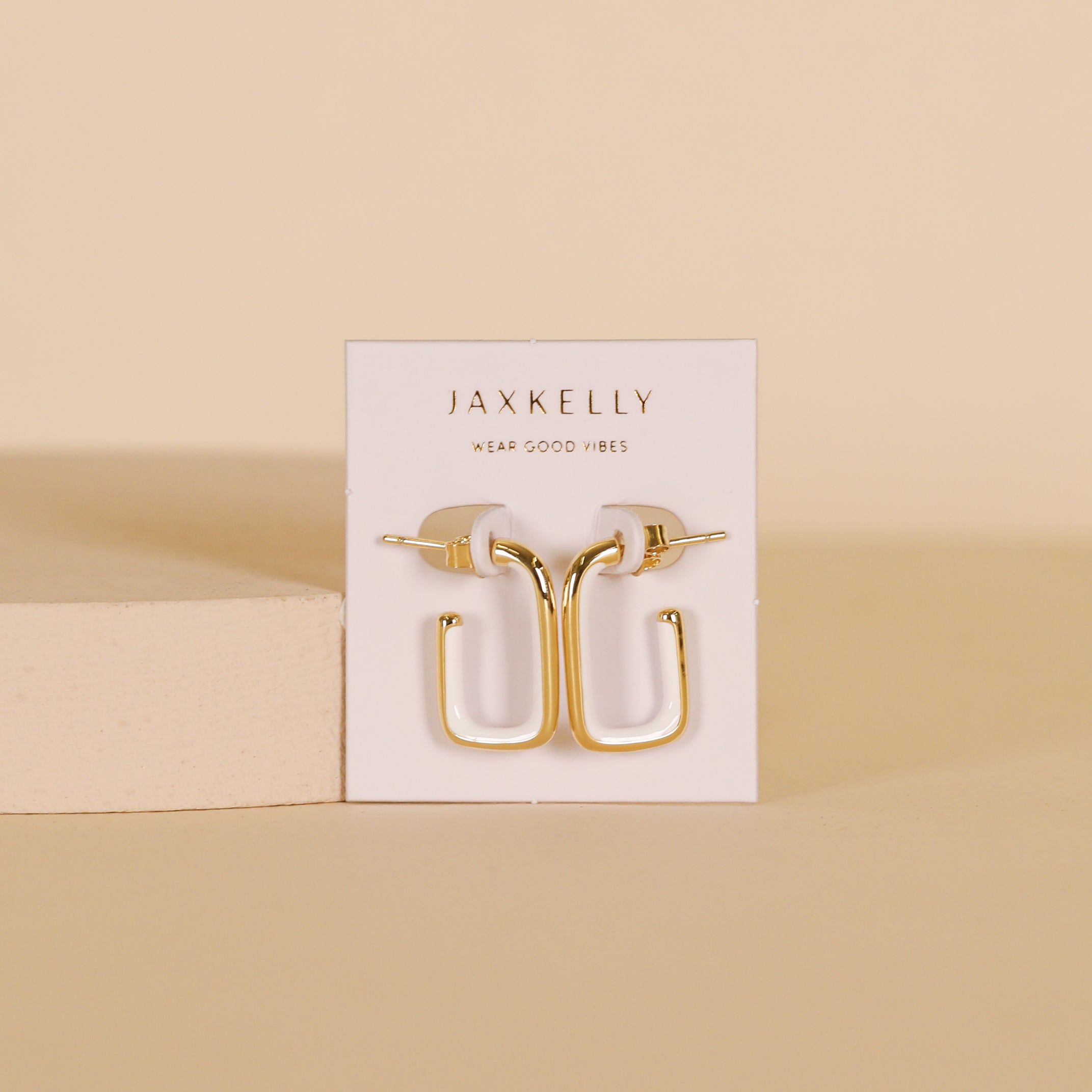 The White Color Dipped Large Rectangle Hoop Earrings are shown on a white JaxKelly card, standing upright against a beige background with a light rectangular block beside them.