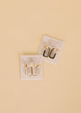Two pairs of JaxKelly earrings on display cards: one pair is 18k gold plated hoops, the other is the White Color Dipped Large Rectangle Hoop Earring, with gold and white enamel accents, both set against a light beige background.