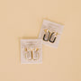 Two pairs of JaxKelly Black Enamel Large Rectangle Hoop Earrings with sterling silver posts rest on branded cards; one pair is all gold, the other features a half gold, half hand-painted black enamel design on a beige surface.