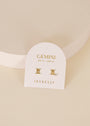 A white card labeled "Gemini May 21 - June 20" and "JaxKelly" displays Zodiac Earrings - Gemini: two radiant 18k gold-plated studs, one shaped like the Gemini symbol and the other accented with a clear gem, on a neutral background.