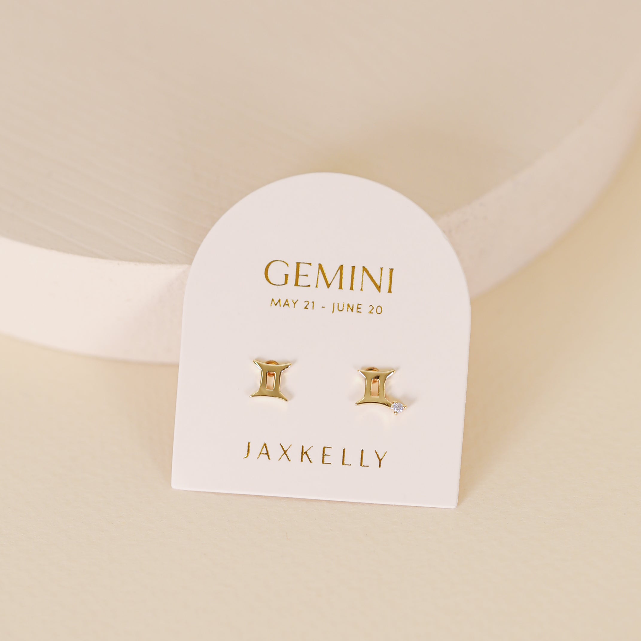 A white card labeled "Gemini May 21 - June 20" and "JaxKelly" displays Zodiac Earrings - Gemini: two radiant 18k gold-plated studs, one shaped like the Gemini symbol and the other accented with a clear gem, on a neutral background.