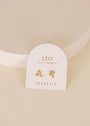 A pair of Zodiac Earrings - Leo by JaxKelly, 18k gold plated, on a white card labeled “LEO July 23 - August 22.” One stud has the Leo symbol; the other is a star with a gemstone accent.