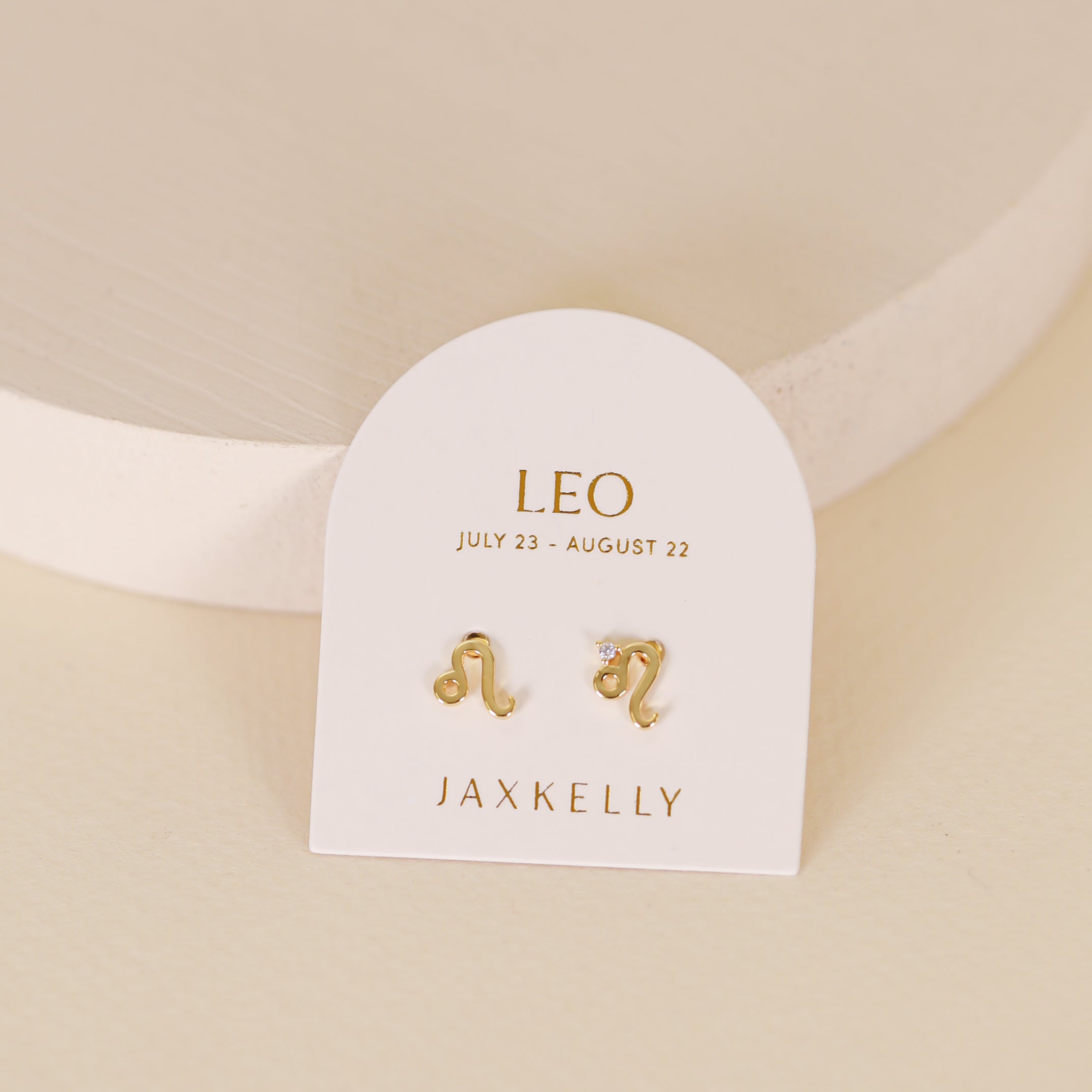 A pair of Zodiac Earrings - Leo by JaxKelly, 18k gold plated, on a white card labeled “LEO July 23 - August 22.” One stud has the Leo symbol; the other is a star with a gemstone accent.