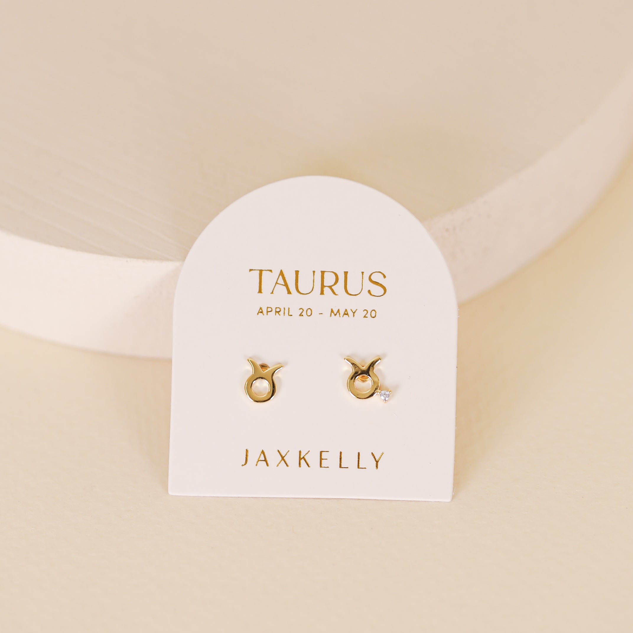 A pair of Zodiac Earrings - Taurus, gold plated studs on a white card labeled "Taurus, April 20 - May 20, JaxKelly," set against a light beige background—an ideal gift for any zodiac jewelry lover.