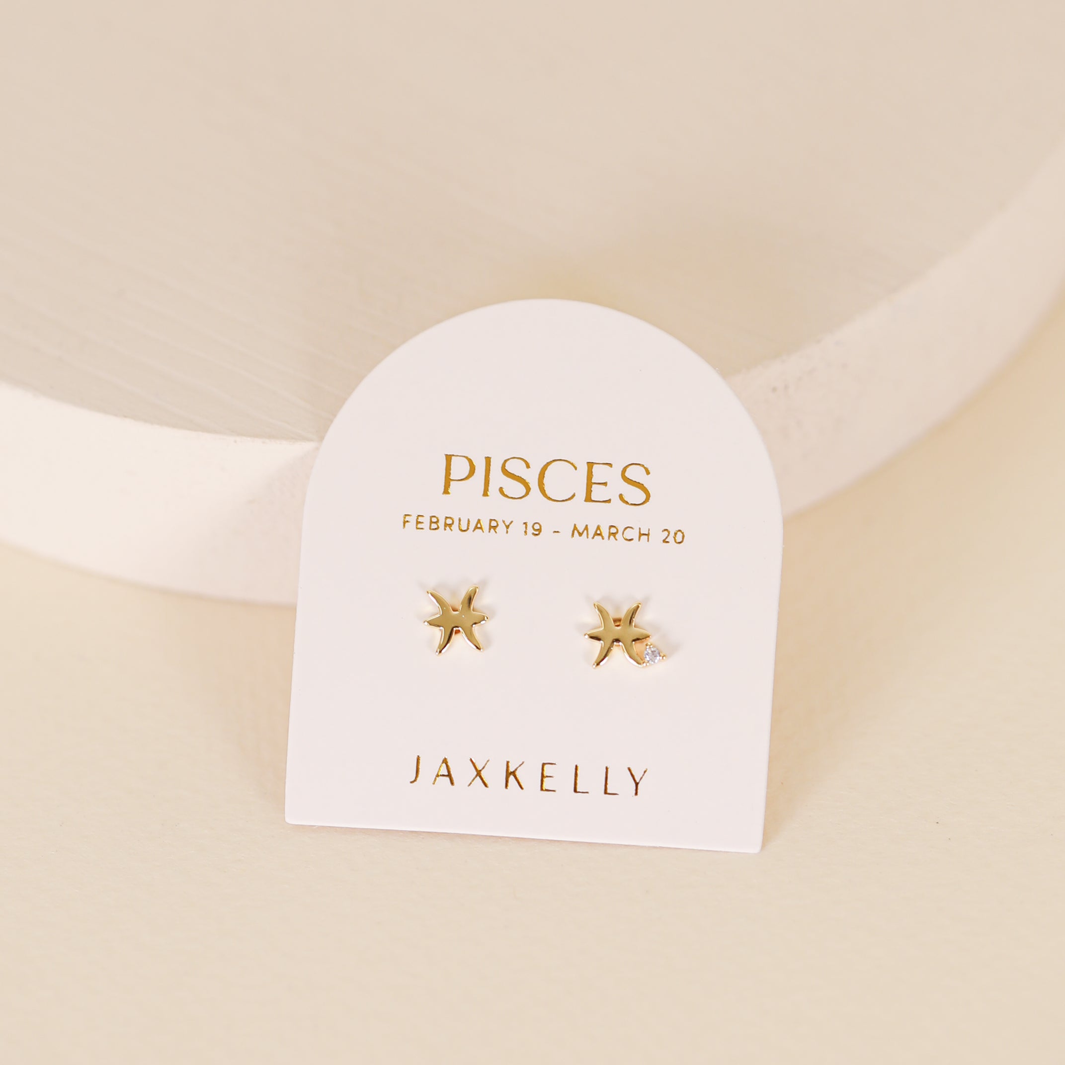 The Zodiac Earrings - Pisces feature small 18k gold plated star-shaped studs on a white card reading "PISCES February 19 - March 20" and "JAXKELLY" in gold, set against a soft beige background.