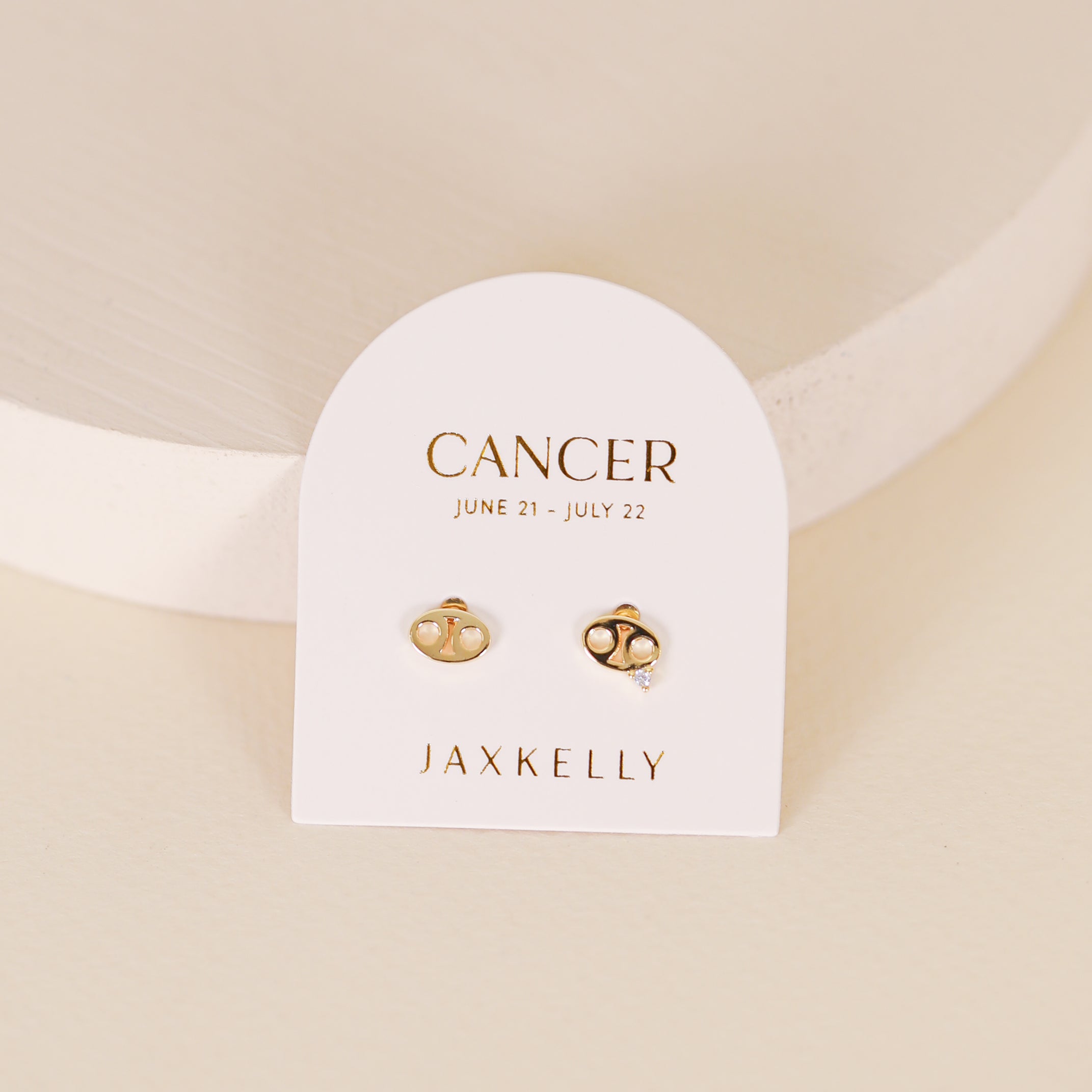 The Zodiac Earrings - Cancer are gold plated studs displayed on a white card reading “CANCER June 21 - July 22” and “JAXKELLY” set on a minimal, neutral-toned backdrop—an elegant choice for any ear piercings collection.