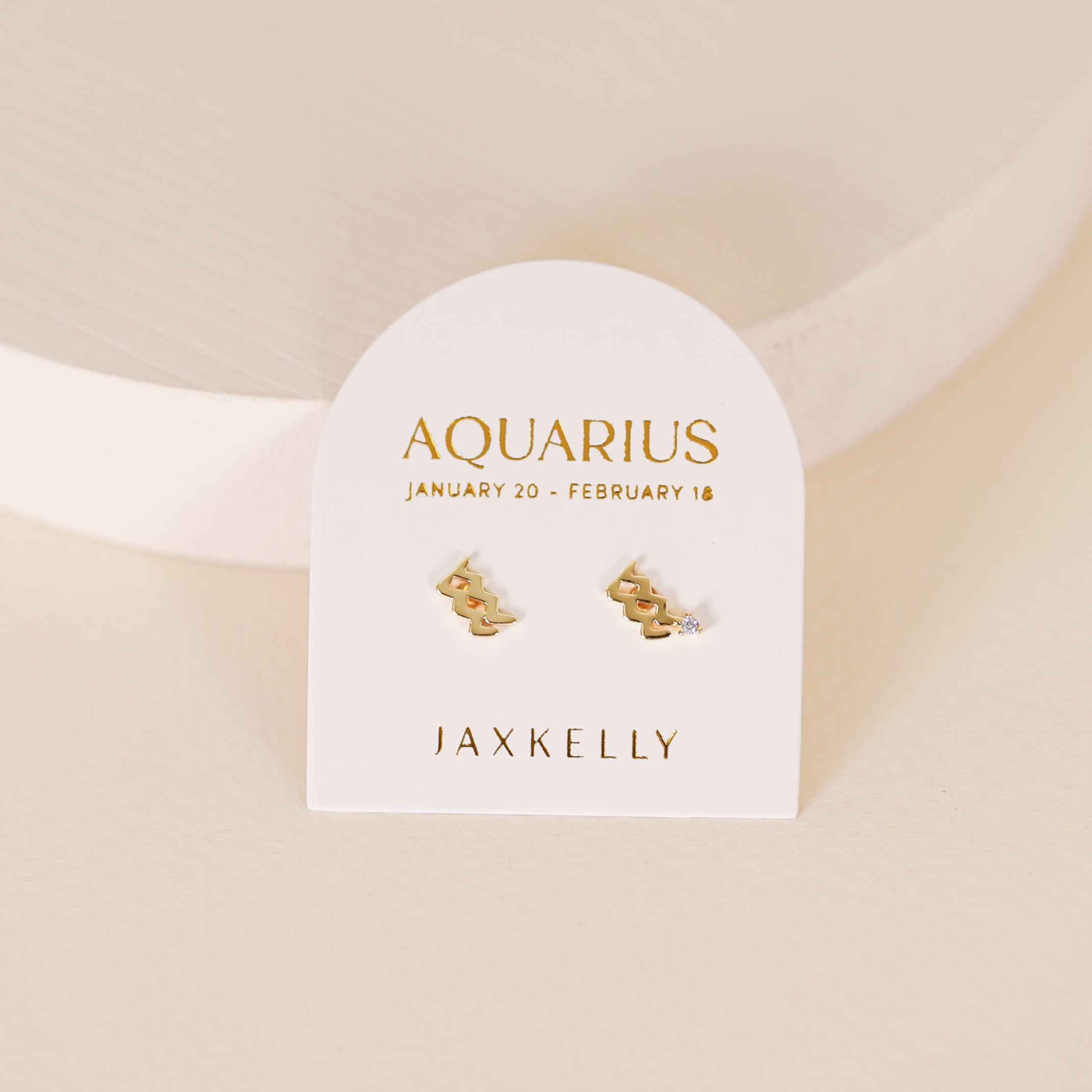 The Zodiac Earrings - Aquarius, featuring the Aquarius symbol in 18k Gold Plating, are shown on a white AQUARIUS JANUARY 20 - FEBRUARY 18 JAXKELLY card atop a textured surface—ideal for ear piercings.