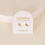 The Zodiac Earrings - Aquarius, featuring the Aquarius symbol in 18k Gold Plating, are shown on a white AQUARIUS JANUARY 20 - FEBRUARY 18 JAXKELLY card atop a textured surface—ideal for ear piercings.