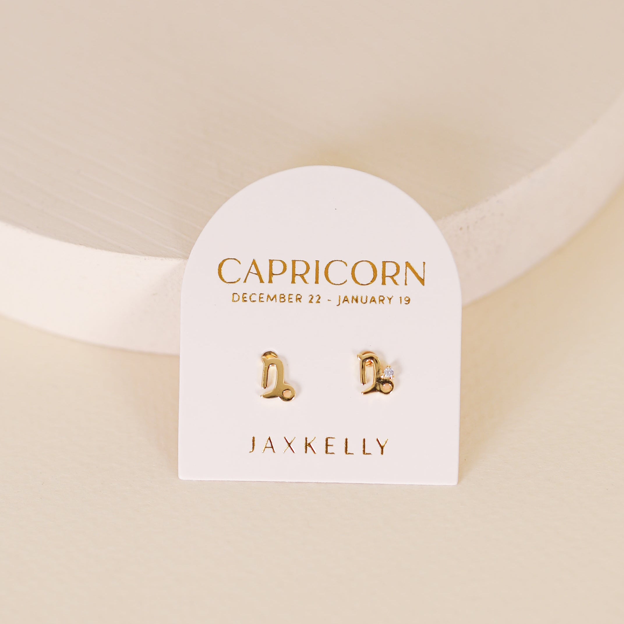 Zodiac Earrings - Capricorn: 18k Gold Plated studs shaped like the Capricorn symbol, displayed on a white card labeled "Capricorn December 22 - January 19" and "JaxKelly"—perfect for ear piercings.
