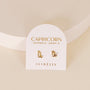 Zodiac Earrings - Capricorn: 18k Gold Plated studs shaped like the Capricorn symbol, displayed on a white card labeled "Capricorn December 22 - January 19" and "JaxKelly"—perfect for ear piercings.