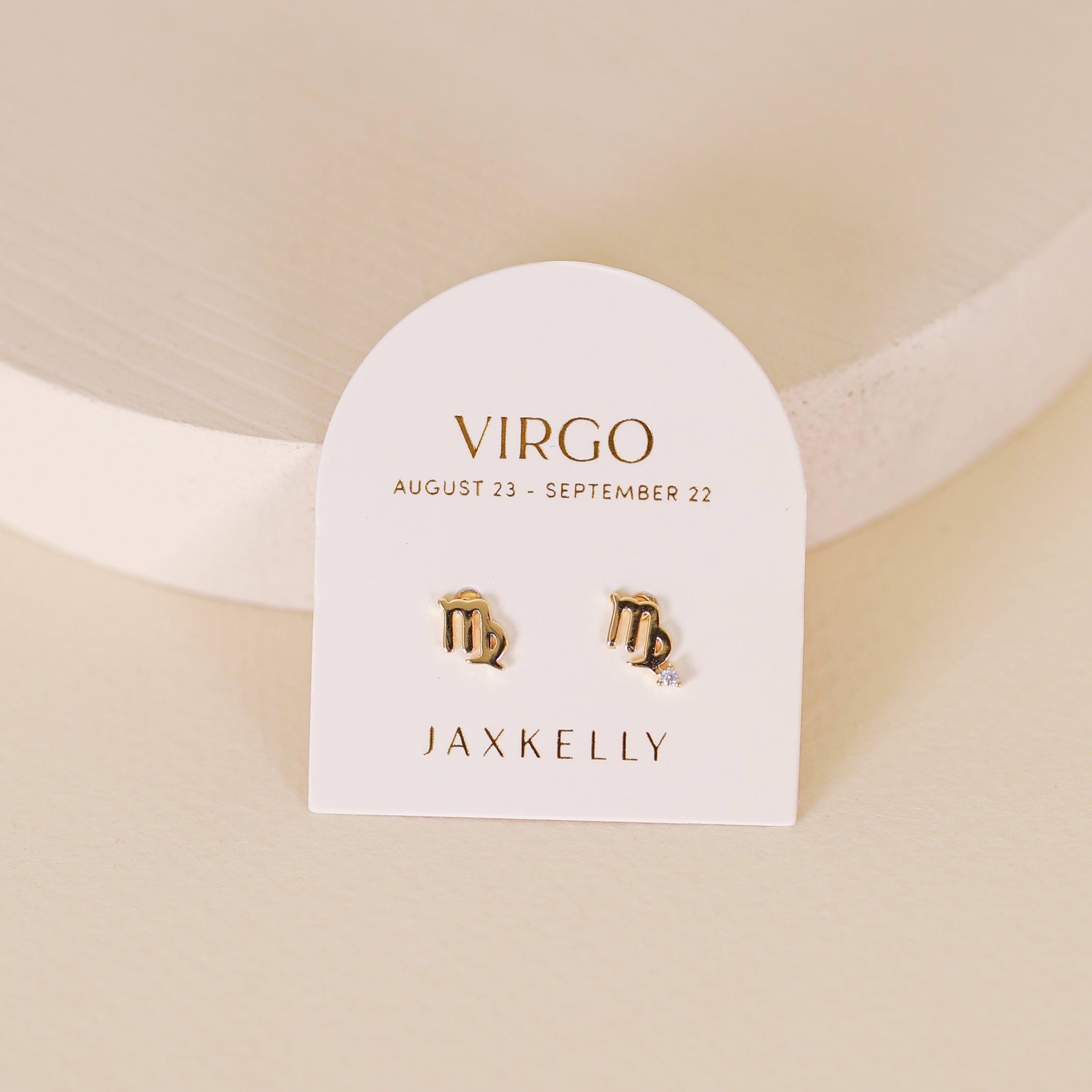 The Zodiac Earrings - Virgo, plated in 18k gold, are displayed on a white card stating “Virgo, August 23 – September 22, JaxKelly” and propped upright on a cream-colored surface.