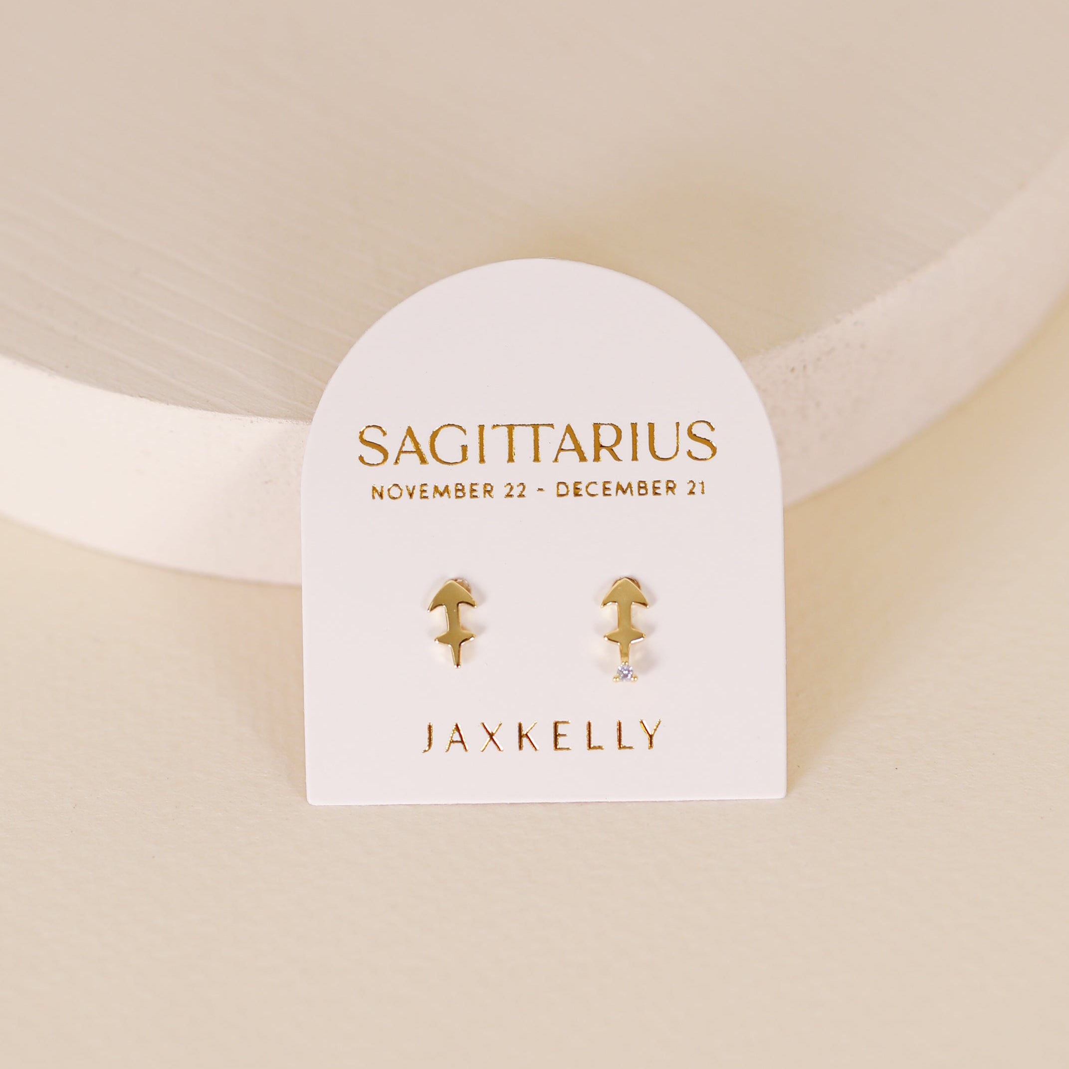 A pair of 18k Gold Plating arrow-shaped Zodiac Earrings - Sagittarius displayed on a white card labeled SAGITTARIUS November 22 - December 21, with the JAXKELLY brand name in gold; set against a softly lit, neutral background.