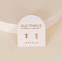 A pair of 18k Gold Plating arrow-shaped Zodiac Earrings - Sagittarius displayed on a white card labeled SAGITTARIUS November 22 - December 21, with the JAXKELLY brand name in gold; set against a softly lit, neutral background.