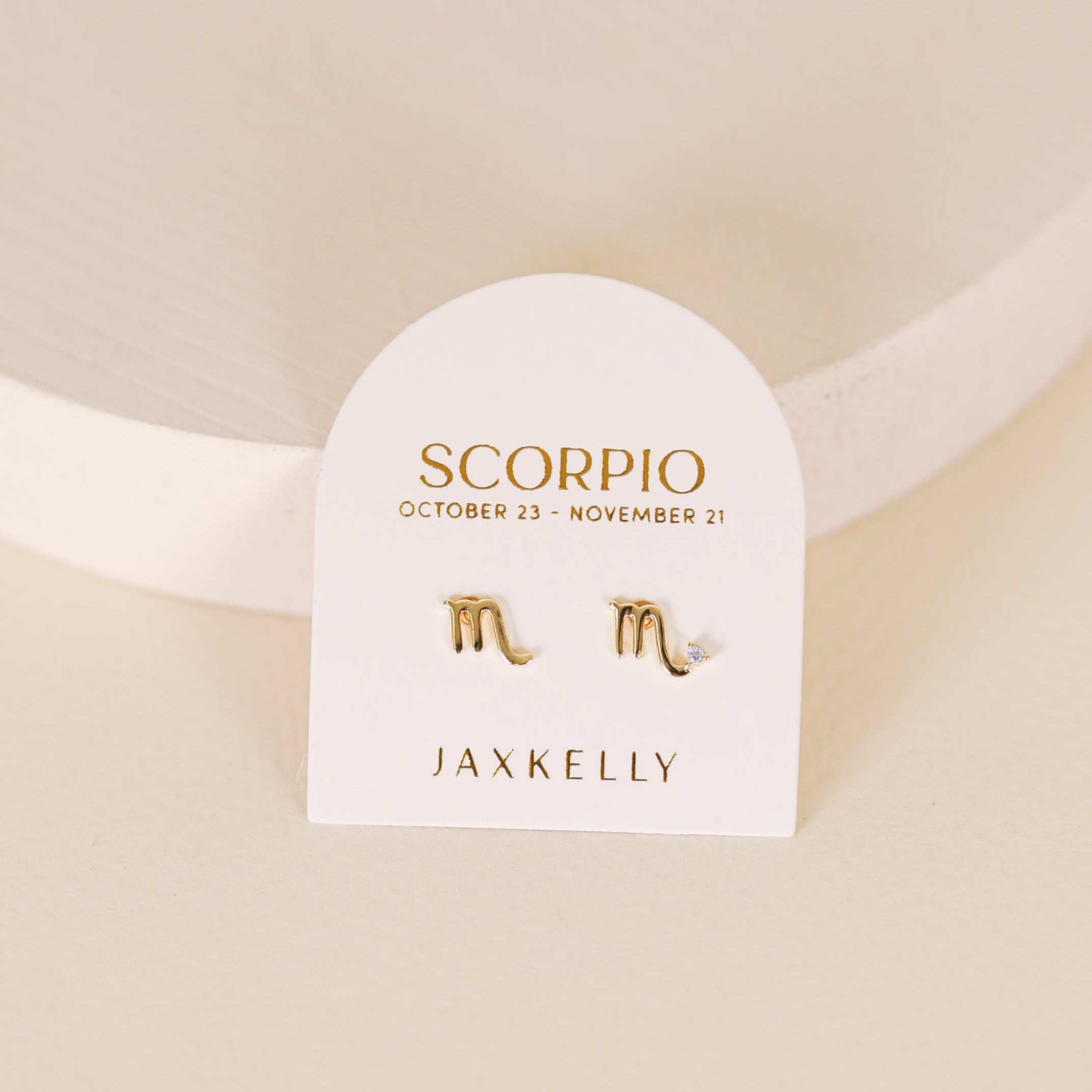 The Zodiac Earrings - Scorpio are gold-plated studs with Scorpio symbols, shown on a white card labeled "SCORPIO October 23 - November 21" and "JAX KELLY." Perfect for ear piercings, they are displayed against a light neutral background.
