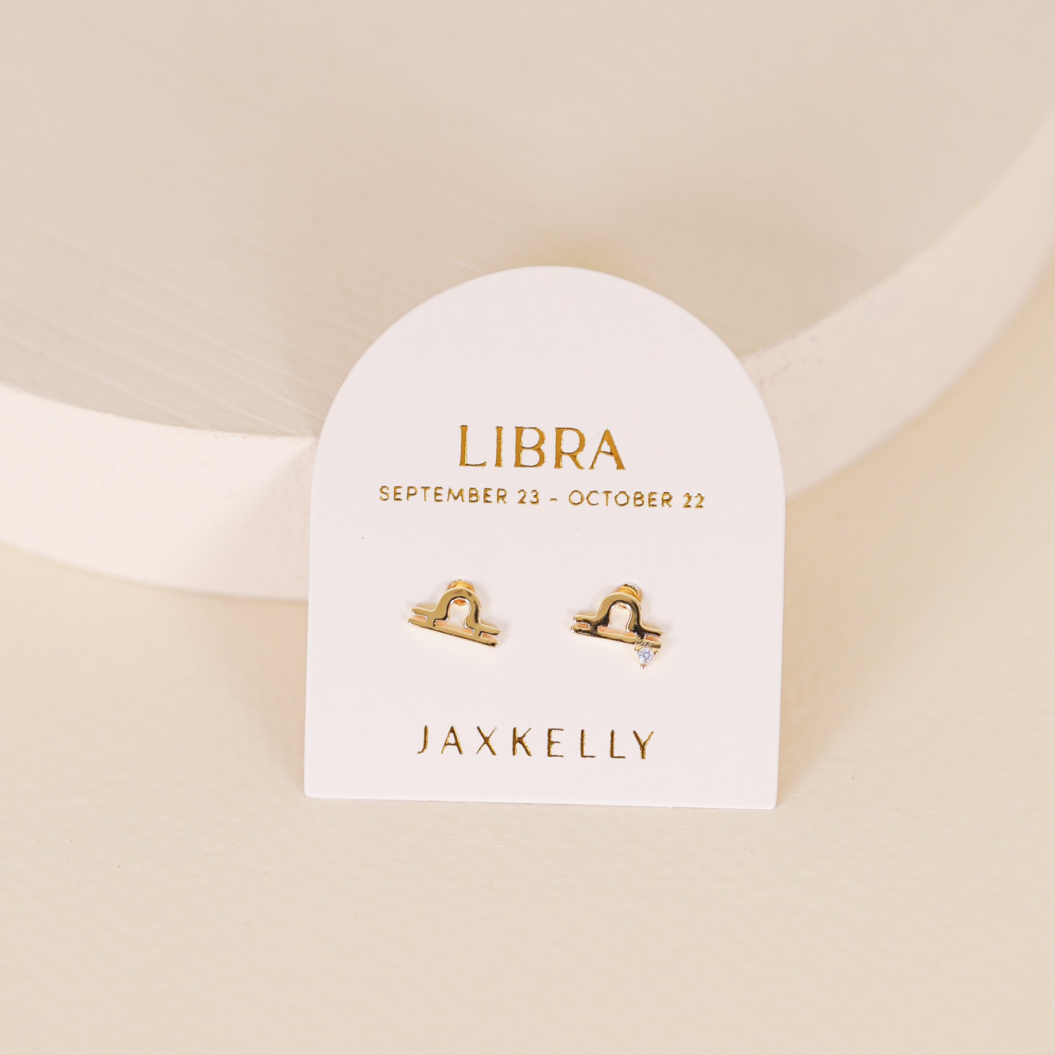 The Zodiac Earrings - Libra are 18k gold plated studs shaped like the Libra zodiac sign, shown on a white card labeled "LIBRA SEPTEMBER 23 - OCTOBER 22 JAXKELLY," set against a neutral background.