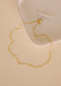The Gold Ball Necklace, featuring 18k gold plating and delicate bead accents, is displayed in a loose circle on a cream surface near a white ceramic plate. The adjustable chain clasp and small tag are shown at the top of the necklace.