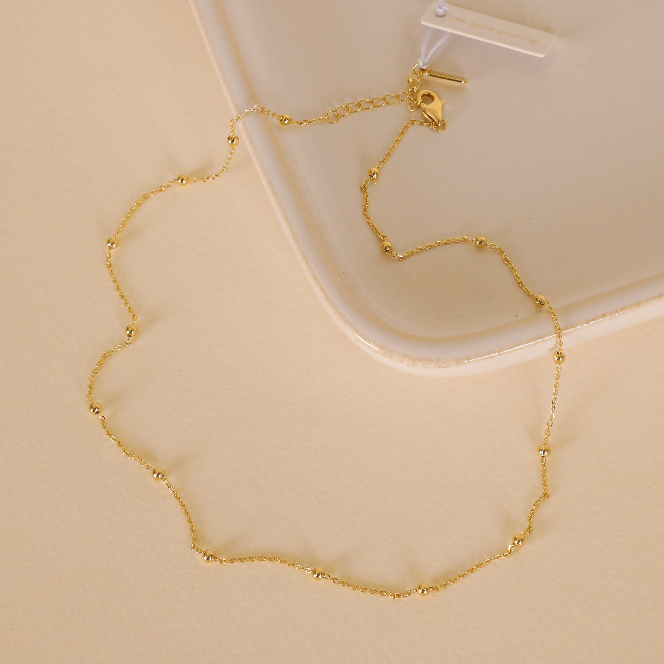 The Gold Ball Necklace, featuring 18k gold plating and delicate bead accents, is displayed in a loose circle on a cream surface near a white ceramic plate. The adjustable chain clasp and small tag are shown at the top of the necklace.