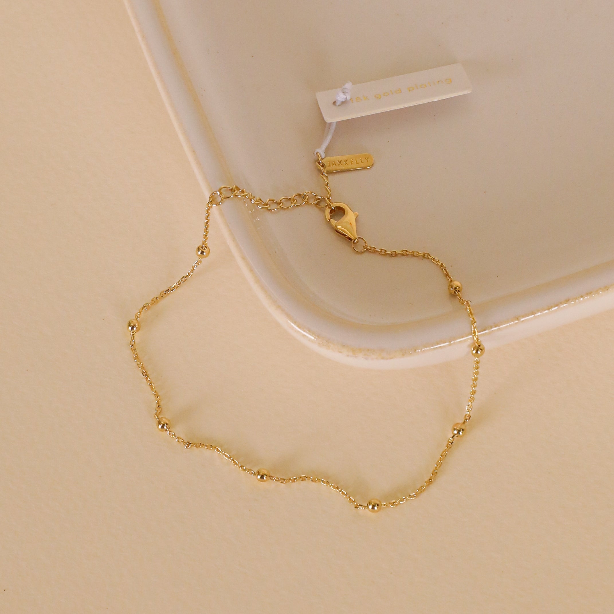The Gold Ball Bracelet, featuring 18k gold plating, small beads, and an adjustable clasp, is displayed on a light beige textured surface next to an off-white ceramic dish. A tag is attached to the bracelet.