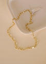 The Large Link Chain Necklace, featuring rectangular links and 18k gold plating, is shown on a beige ceramic tray with a matching backdrop. A white tag on one end highlights its adjustable chain design.
