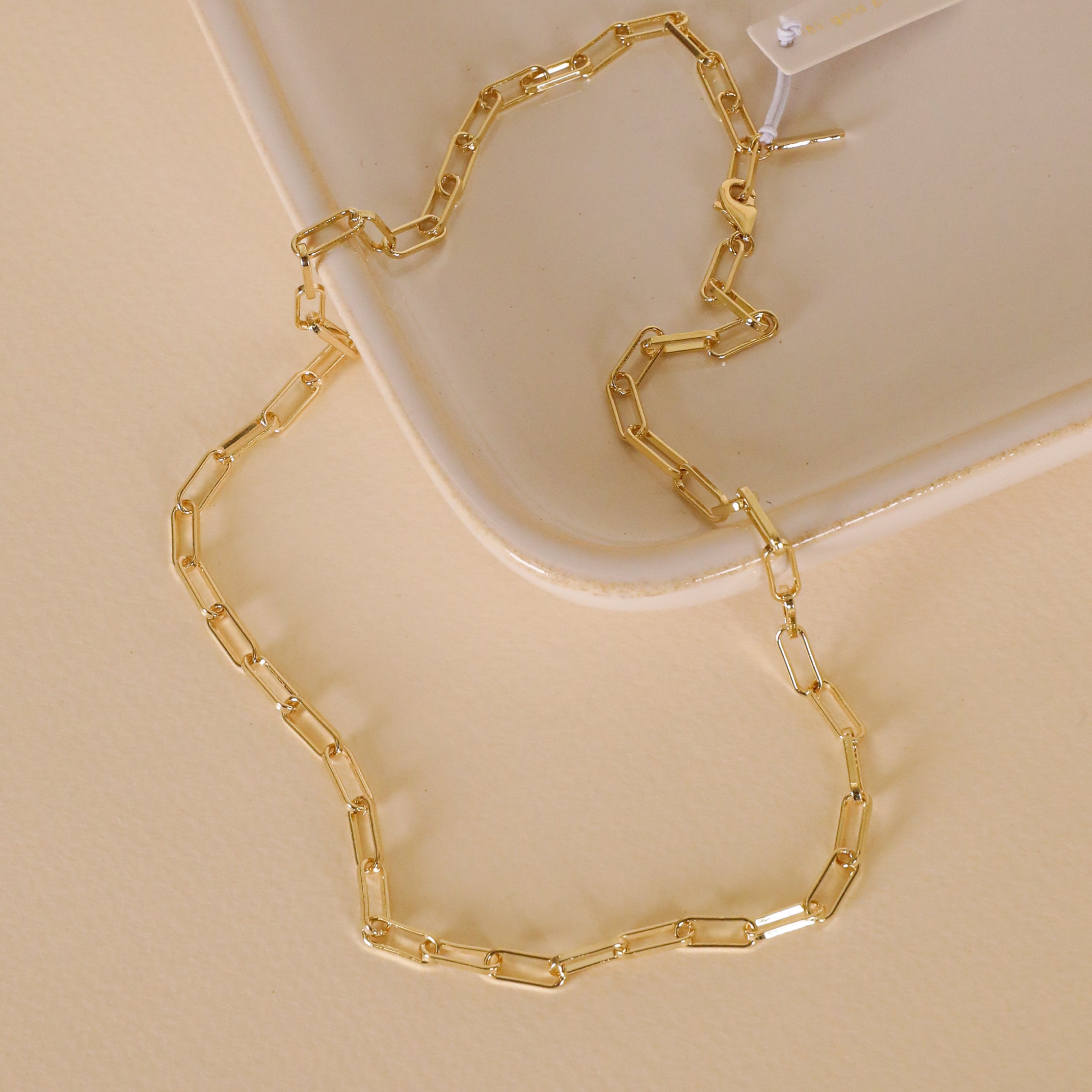 The Large Link Chain Necklace, featuring rectangular links and 18k gold plating, is shown on a beige ceramic tray with a matching backdrop. A white tag on one end highlights its adjustable chain design.