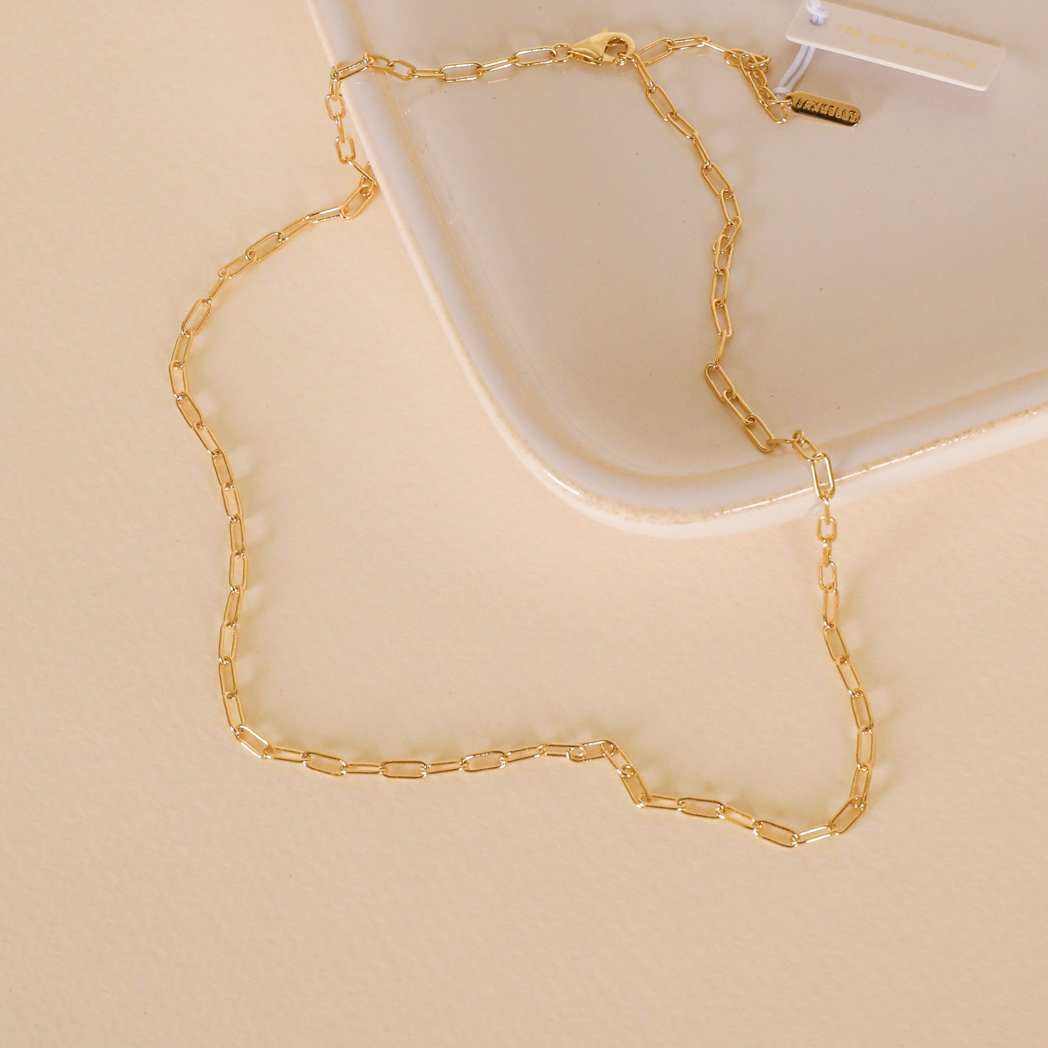 The Small Link Chain Necklace, a delicate gold plated piece with rectangular links, is displayed on a cream-colored surface beside the corner of a square, off-white tray.