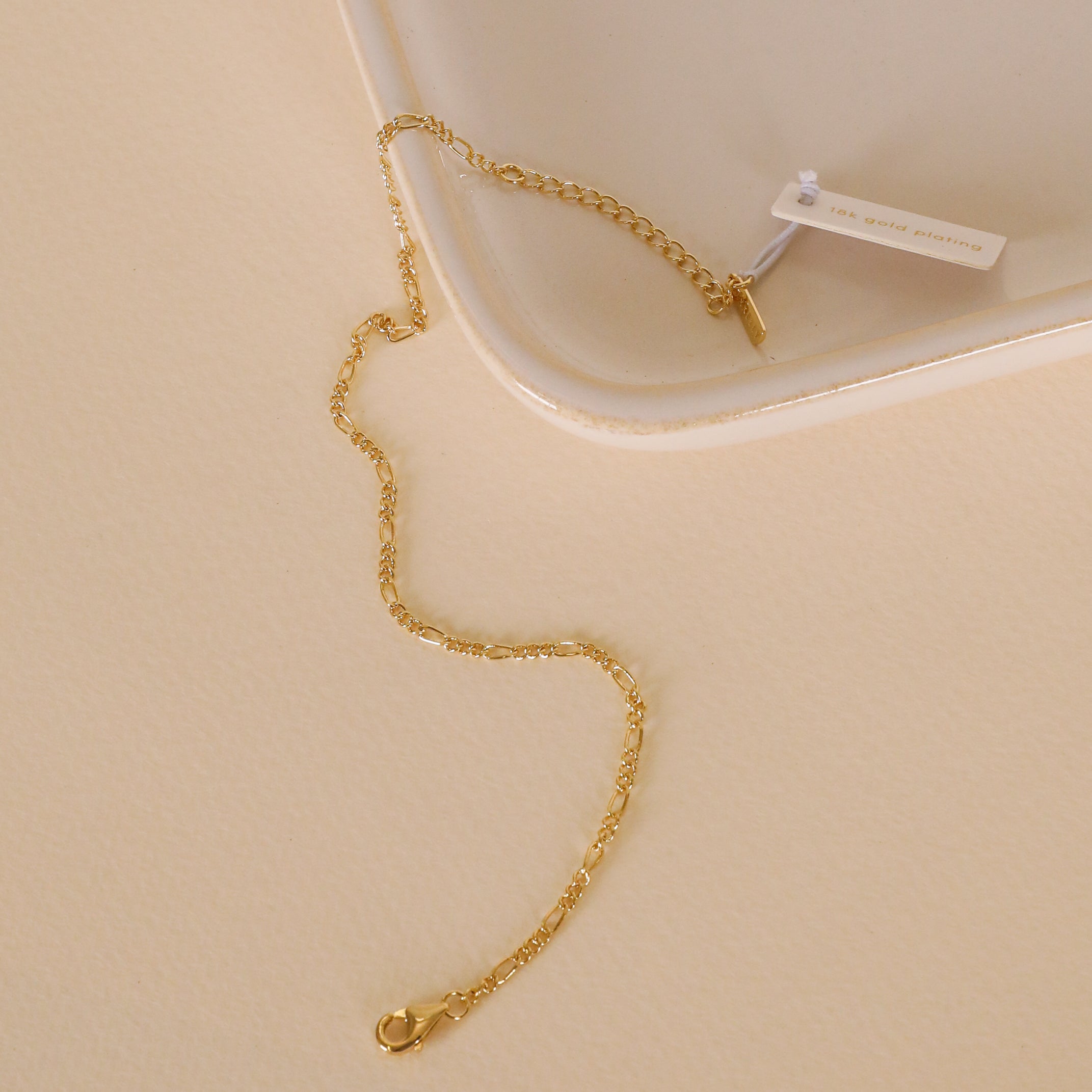 The Simple Twist Chain Bracelet by JaxKelly, featuring a delicate 18K gold plated design with a small tag, rests on a light beige surface beside the corner of a white tray. Its tarnish-free shine ensures lasting beauty for everyday wear.