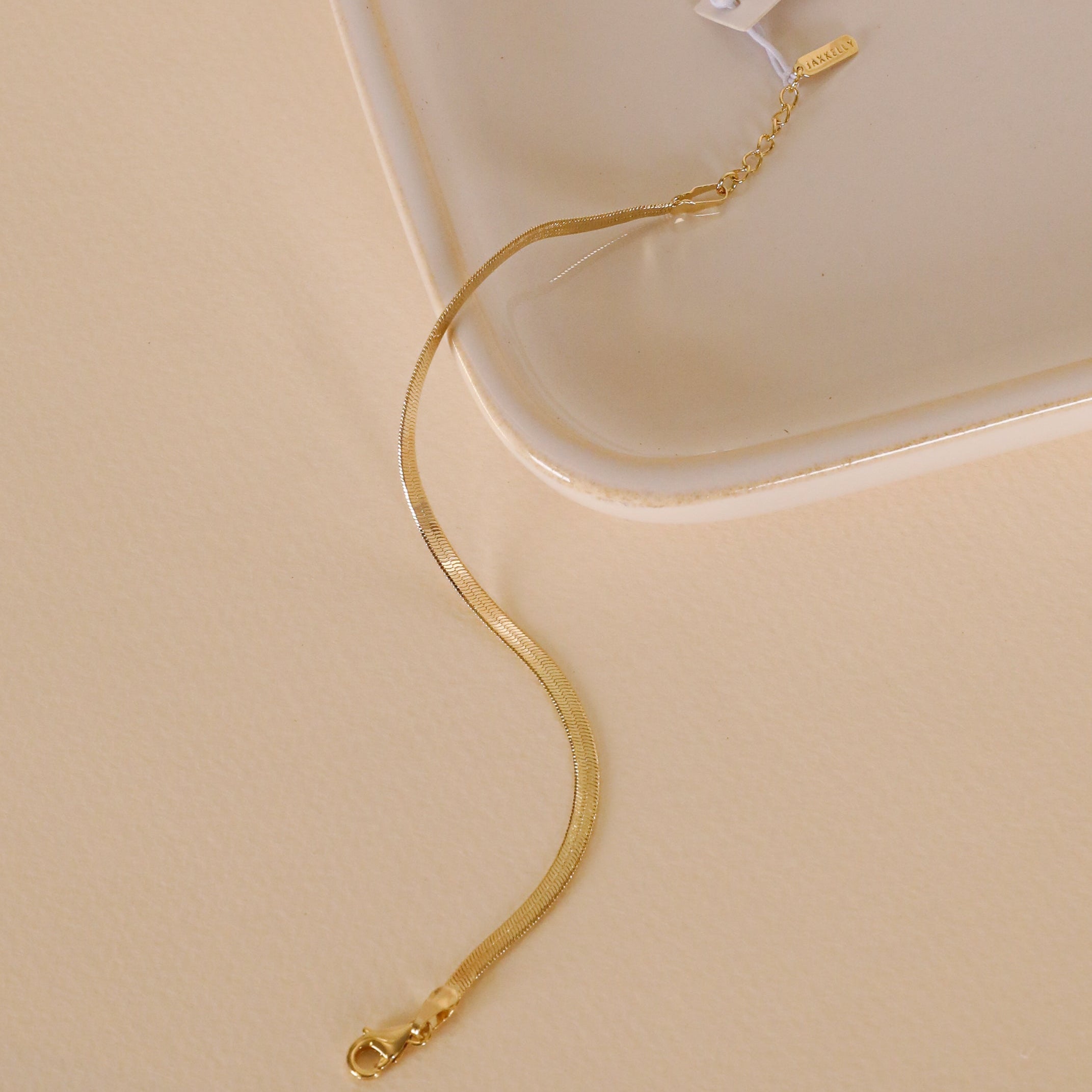 The Flat Snake Chain Bracelet, featuring an 18k gold-plated delicate snake chain and a lobster clasp, rests on a light cream surface beside a ceramic tray.