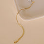 The Flat Snake Chain Bracelet, featuring an 18k gold-plated delicate snake chain and a lobster clasp, rests on a light cream surface beside a ceramic tray.