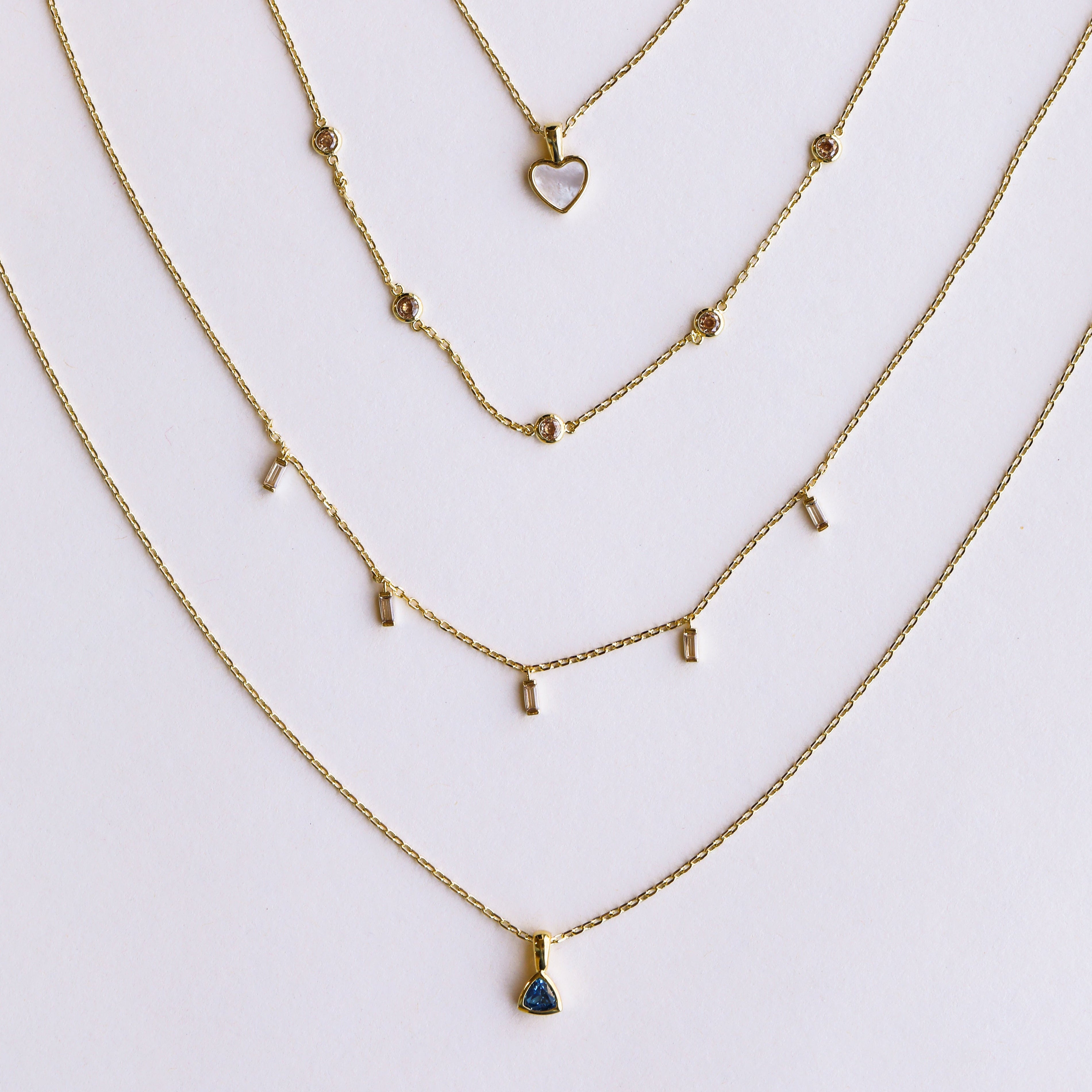 Four delicate gold necklaces by JaxKelly are arranged in a semi-circle. Discover the Mystery Necklace among heart, circles, bars, and blue triangle stone pendants—perfect for jewelry sales or as a surprise gift.